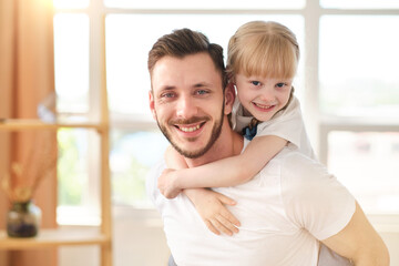 Fototapeta premium Attractive young father standing in the living room holds on his back a charming daughter at home. Smiling daughter spends time with a loving father, playing a piggyback with dad. Father's day.