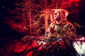 Medieval monk in canvas sackcloth robe praying in dark forest with snow and red light on winter...