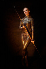 Valkyrie girl in shiny military armor and with a spear in a dark room with plants and vines. Model...