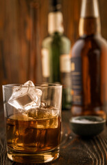 A glass of whiskey with ice, in the background are bottles on a wooden table of a bar counter, shallow depth of field, selective focus. The concept of alcoholic drinks in a roadside bar.