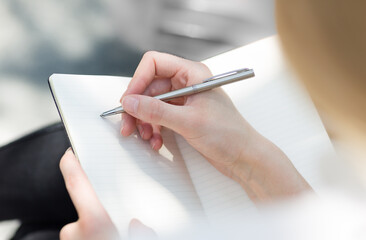 Closeup of a female's hand writing on an blank notebook with a pen sitting outdoors in the sun. Unrecognizable crop. 