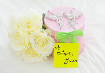 Valentines day romantic gift: bouquet of white roses, gift box with diamond earrings and a "I Love You" note placed on the bed.