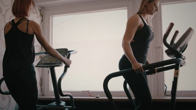 Two fit women exercise on cardio machines in gym, slow motion