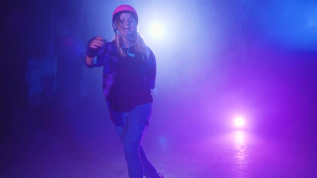 Happy and cheerful girl, a professional roller in a helmet and protection, dances and performs U-turns under the bright lighting of blue and pink in the smoke of an abandoned platform on the stage.