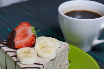 Cheesecake on a plate. Garnished with strawberries, bananas and chocolate. Near a cup of coffee. On brushed pine boards painted black and green.