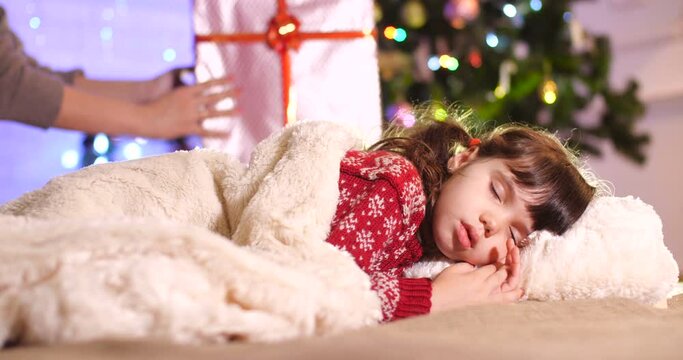   Little European brunette  girl in a burgundy sweater with a pattern sleeping  behind a white fur blanket near the Christmas tree. In the background, mom&rsquo;s hands are laying a box with a gift. 
