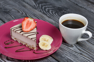 Cheesecake on a plate. Garnished with strawberries, bananas and chocolate. Near a cup of coffee. On brushed pine boards painted in black and white.