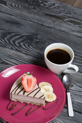 Cheesecake on a plate. Garnished with strawberries, bananas and chocolate. Near a cup of coffee. On brushed pine boards painted in black and white.