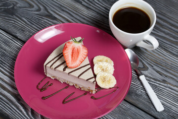 Cheesecake on a plate. Garnished with strawberries, bananas and chocolate. Near a cup of coffee. On brushed pine boards painted in black and white.