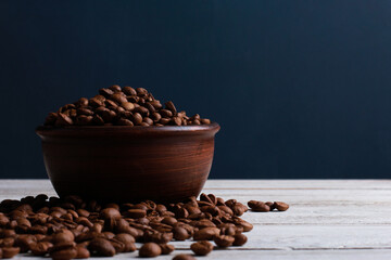 Fototapeta premium Coffee beans in clay bowl on white wooden table against dark blue wall background. Closeup, copy space, side view. Arabica seeds, roast, caffeine concept