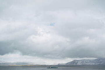 The barge sails on the Atlantic Ocean, near Iceland, against the backdrop of snow-capped mountains. Gloomy cloudy sky.