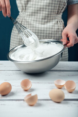 Woman in checkered apron whips egg whites on a white vintage wooden table.
