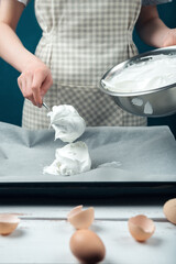 Woman spreads beaten egg whites on a baking sheet covered with parchment on a white vintage wooden table.