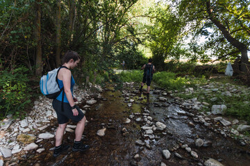 Naklejka na ściany i meble Small group of young tourists walking inside a river with little flow inside a forest in Spain in spring. Selective focus.