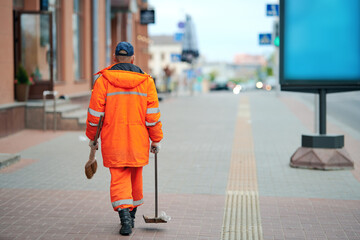 Municipal worker in orange uniform collecting garbage from pedestrian street and footpath. City...