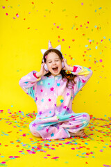 Obraz na płótnie Canvas little girl with long hair wearing a kigurumi and headphones in the shape of a unicorn is happy catching confetti standing on a yellow background