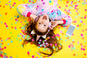 Fototapeta premium happy little girl in a unicorn kigurumi lies on a yellow background amid multicolored confetti listening to music