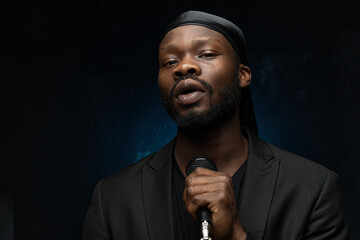 dark-skinned handsome guy in a bandana, black classic jacket and t-shirt holds a microphone in his hand, looks in the camera and singing