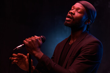 dark-skinned handsome guy in a bandana, black classic jacket and t-shirt holds a microphone in his hands and emotionally sings in a dark studio with red light