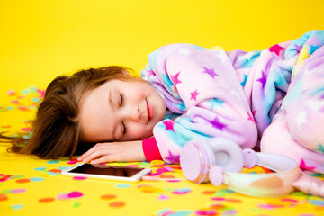 happy little girl in a unicorn kigurumi lies on a yellow background amid multicolored confetti listening to music