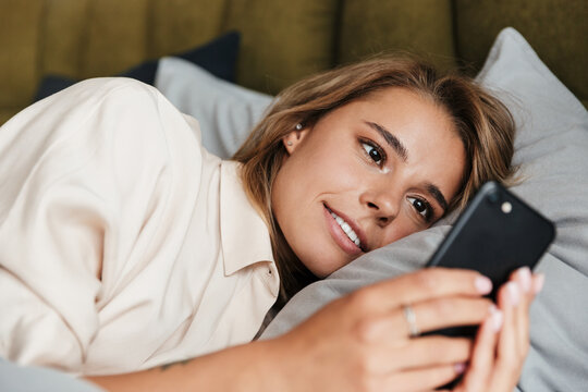 Image of pleased woman smiling and using cellphone while lying in bed