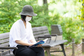 Fototapeta premium 公園のベンチで帽子をかぶりマスクをして読書する1人の女性