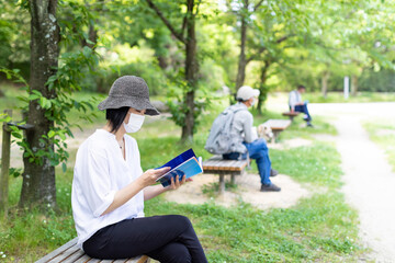 公園のベンチで帽子をかぶりマスクをして読書する1人の女性