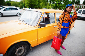 Beautiful african american lady with shopping bags standing near orange classic retro car. - Powered by Adobe