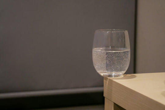 A glass of water with bubbles inside is on the corner of light yellow wooden table in the grey room. A cup on the edge looks dangerously.