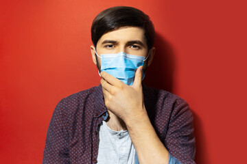 Studio portrait of young guy wearing medical face mask, protection against coronavirus. Prevention of covid-19. Background of red color.