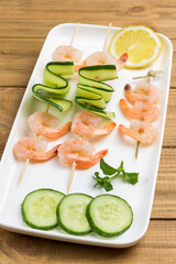 Shrimps with cucumber and tomatoes on skewer in white plate.