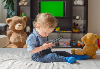 Cute baby boy playing with the remote control to watch TV sitting on a couch with his teddy bear,...