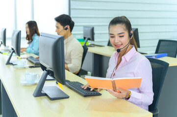 Customer support representative talking on headset while using computer. Businessman working at desk with colleague in background. He is wearing casuals in call center.