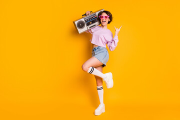 Full length body size view of nice attractive funny cheerful cheery excited glad girl carrying boombox having fun dancing showing horn sign isolated bright vivid shine vibrant yellow color background