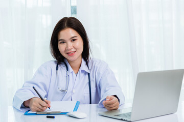 Fototapeta na wymiar Asian doctor young beautiful woman smiling using working with a laptop computer and her writing something on paperwork or clipboard white paper at hospital desk office, Healthcare medical concept