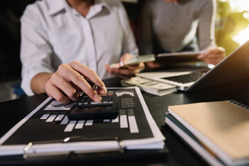 Business documents on office table with smart phone and laptop and two colleagues discussing data in the background in morning light
