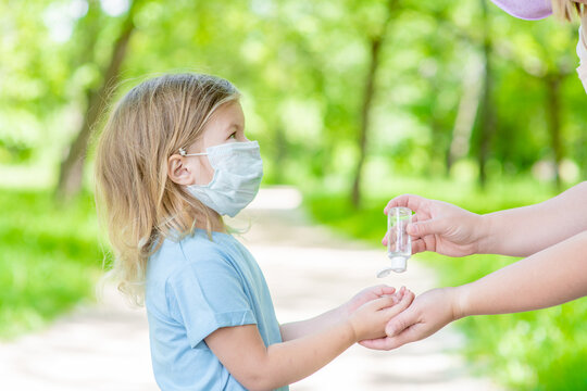 Mother applies sanitizer for cleaning baby hands in public place. Little girl wearing protective face mask looks at mom