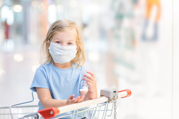 Little girl wearing protective facemask during coronavirus and flu outbreak sits inside shopping cart and applies sanitizer for cleaning hands in public crowded place