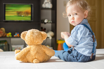 Cute baby boy and his teddy bear watching TV sitting on a couch in the living room at home