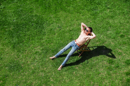 A Man Sunbathes On A Chair With A Naked Torso And Barefoot On A Background Of Green Grass. Man Resting In The Park On The Lawn