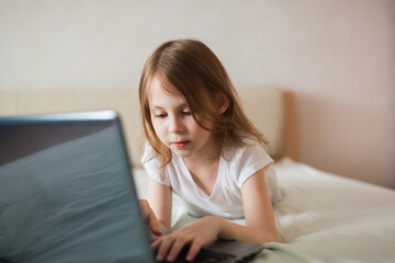 Close-up portrait of a baby girl with a laptop on the bed. The concept of digital communication, digital addiction, entertainment, distance learning online.