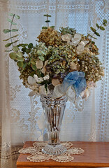 This vertical still life shows dried hydrangea and other florals in a crystal vase, on top of...