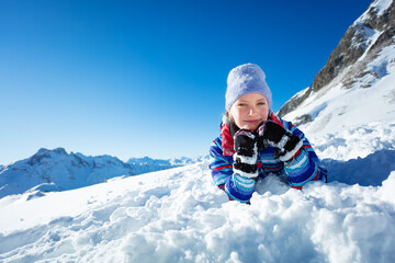 Fototapeta na wymiar Happy smiling little girl lay in snow laying putting head on hands and smile wearing winter hat with mountain on background