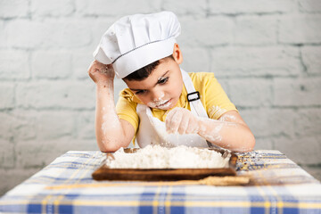 un niño vestido con gorro y delantal de panadero agarra harina con la mano, tiene la boca y cara...