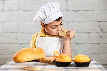 a boy wearing a hat and a baker's apron takes a slice of bread to his mouth while he bites and eats it and with the other hand holds another piece. rustic scene. lifestyle, bakery.