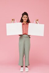 Portrait of young woman with curly hair smiling at camera and holding two shopping bags in her hands against the pink background