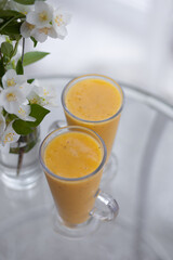 two yellow smoothie cocktails in high glasses on a glass breakfast table in the morning with white flowers. Side view. Summer Spring