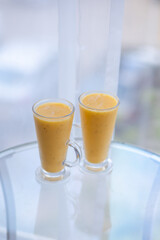 two yellow smoothie cocktails in high glasses on a glass breakfast table in the morning with white flowers. Side view. Summer Spring