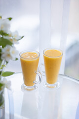 two yellow smoothie cocktails in high glasses on a glass breakfast table in the morning with white flowers. Side view. Summer Spring