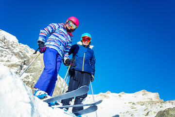 Two girls stand on top of the mountain on snow pile, view from below with ski and skiers equipment
